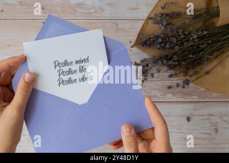 Weibliche Hände, die eine Notiz aus Papier nehmen, mit Text POSITIVE MIND LIFE VIBES aus violettem Umschlag. Lavendelblume. Draufsicht, flach. Konzept der psychisch-spirituellen Gesundheit Selbstversorgung Wohlbefinden Achtsamkeit Stockfoto