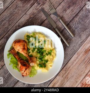Ein Teller mit zwei gebackenen Hähnchenschenkeln und Kartoffelpüree auf einem Holztisch mit Besteck. Draufsicht, flach. Stockfoto