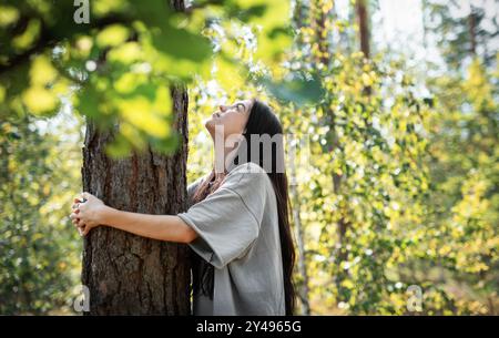 Eine Frau lehnt sich an einen Baum, blickt mit einem ruhigen Ausdruck nach oben, umgeben von leuchtend grünen Blättern in einem sonnendurchfluteten Wald. Stockfoto