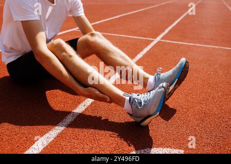 Ein Läufer, der auf einer Schiene sitzt und sein Bein unter Schmerzen hält, möglicherweise aufgrund eines Krämpfes oder einer Verletzung. Stockfoto
