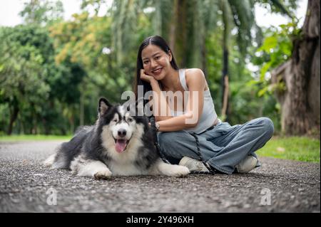 Ein wunderschöner sibirischer Husky-Hund an der Leine und seine asiatische Besitzerin lächeln in die Kamera, während sie zusammen auf einer Straße in einem grünen Park sitzen Stockfoto