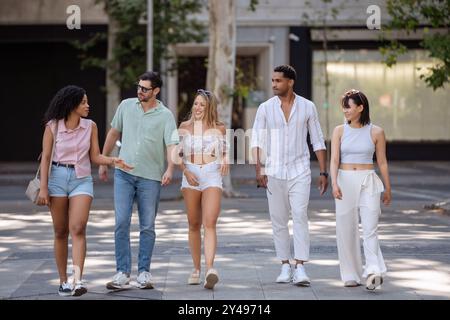 Eine vielfältige Gruppe von fünf jungen Freunden schlendert durch einen Stadtplatz, führt lebhafte Gespräche und genießt einen sonnigen Tag mit Bäumen und urbaner Umgebung. Stockfoto