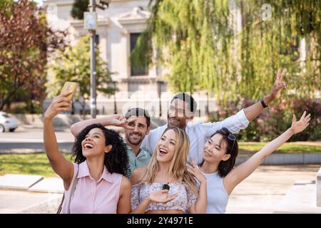 Fünf fröhliche Freunde treffen sich zu einem Selfie in einem sonnigen Park, lächeln, machen Friedensschilder und genießen gemeinsam einen lustigen Moment mit Grün und urbaner Umgebung. Stockfoto