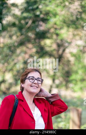 Eine reife Frau mit Brille lächelt warm, während sie einen roten Blazer trägt. Der Hintergrund ist verschwommen, mit üppigem Grün und Sonnenlicht, das durch die Luft filtert Stockfoto