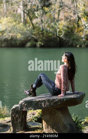 Frau, die die Wintersonne genießt, umgeben von Grün und ruhigem Wasser, sitzt auf einer Steinbank am Fluss Eume in Galicien, Spanien Stockfoto