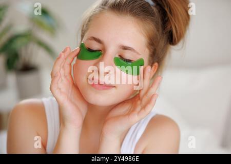 Das Teenager-Mädchen pflegt ihre Haut, trägt grüne, pflegende Flecken unter den Augen auf, vor dem Hintergrund einer häuslichen Umgebung Stockfoto