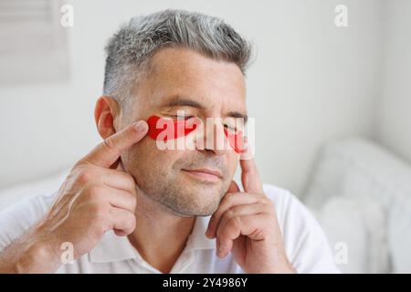 Mann, der Augenflecken auf das Gesicht aufträgt. Falten und Gesichtspflege zu Hause für Männer. Das Konzept der Gesichtsverjüngung Stockfoto