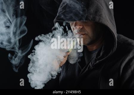 Mann in der Haube, der Dampf ausatmet, umgeben von rauchendem Rauch. Dunkler Hintergrund, elektronisches Zigarettenkonzept Stockfoto