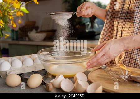 Nahaufnahme von weiblichen Händen, die Mehl gesiebt und Teig in einer durchsichtigen Schüssel vor dem Hintergrund einer Küche zubereiten. Eier in einer Kiste und ein Werkzeug für Mak Stockfoto