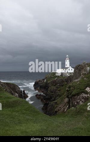 Vertikales Foto des Fanad Head Lighthouse auf einer zerklüfteten Klippe in Irland unter dunklem, stürmischem Himmel, mit Wellen, die gegen die Felsen darunter krachen Stockfoto