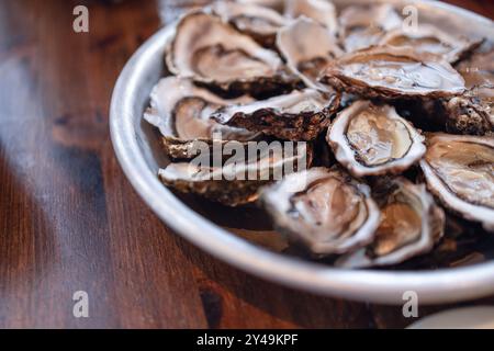 Rohe Austern in geöffneten Schalen füllen ein rundes Metalltablett auf einem Holztisch in Irland, mit glitzerndem Fleisch und strukturierten Schalen in einer Nahaufnahme. Stockfoto