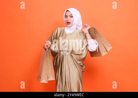 Schockierte junge asiatische Frau, die eine Papiertüte mit Lebensmitteln in einem muslimischen Kaftan-Kleid mit einem Hijab trägt, isoliert auf einem orangen Rücken Stockfoto