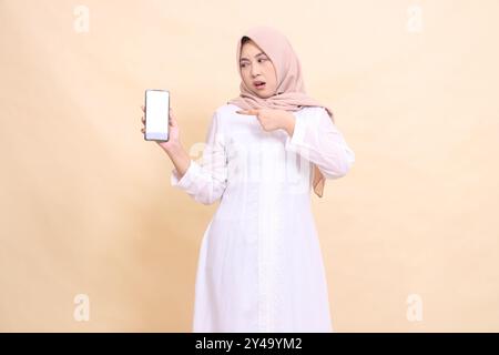 Frau junge asiatische muslime, die einen Hijab trägt und offen schmeckt, während sie ihr Handy-Gerät nach rechts in der Hand hält, während sie zeigt. Lifestyle, Business Stockfoto