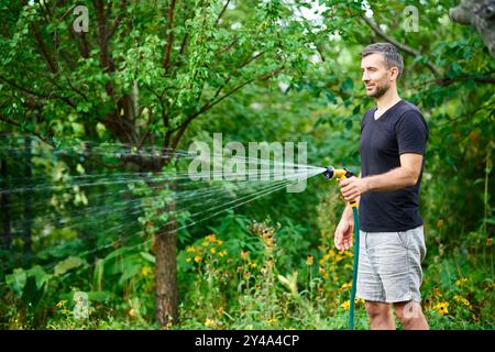 Junger Mann, der Blumen und Pflanzen im Garten mit Schlauch im sonnigen blühenden Hinterhof tränkt Stockfoto
