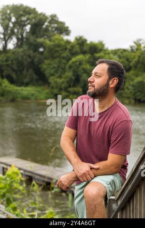 Ein Mann in einem roten Hemd sitzt auf einer Holztreppe mit Blick auf ein Gewässer. Er blickt in die Ferne, verloren in Gedanken. Konzept der Ruhe und Stockfoto