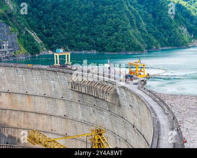 Industriekrane und Ausrüstung auf dem Enguri-Wasserkraftdamm in Georgien. Betonbau kurvt um den Stausee, üppig grüner Wald im Hintergrund, Technik und Natur. Stockfoto
