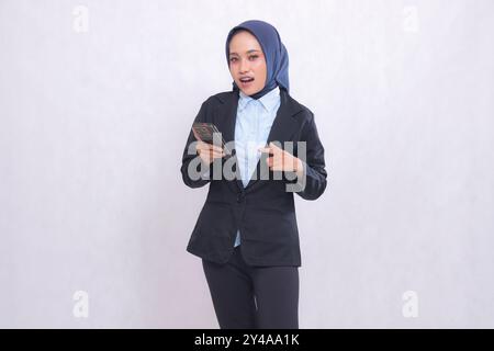 Die asiatische Büroleiterin im Hijab steht überrascht vor der Kamera und zeigt während des Betriebs auf ihr Handy. Schöne muslimische Frau in bl Stockfoto