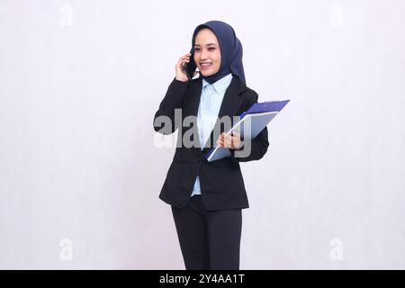 Exklusive asiatische Bürofrau mit Hijab steht, wenn jemand anruft, und trägt einen Stift, ein Klemmbrett und ein Notebook. Schöne muslimische Frau in blauem Hemd Stockfoto