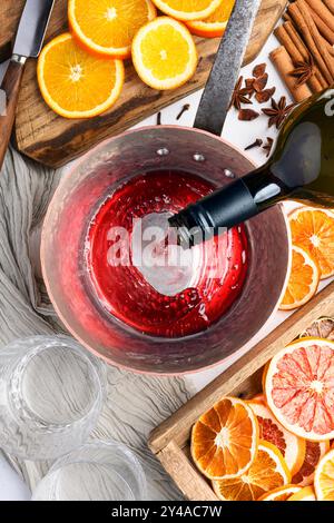 Rotwein, der aus der Flasche in einen Kupfertopf gießt. Glühwein in der Küche zubereiten. Orangenscheiben, Zimt, Sternanis auf dem Tisch. Lebensmittelfotografie Stockfoto