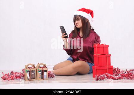 Ein wunderschönes südostasiatisches Mädchen, das bei Weihnachten neben 3 Geschenkboxen mit Hammerboxen mit Weihnachtsschmuck und rotem Hemd auf die Bestellung blickt Stockfoto