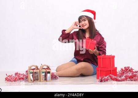 Ein wunderschönes südostasiatisches Mädchen, das mit der Hand lächelt, als Zeichen der Liebe neben 3 Geschenkboxen mit Hammer an Weihnachten mit einem Weihnachtsmann-Haarreif und Stockfoto