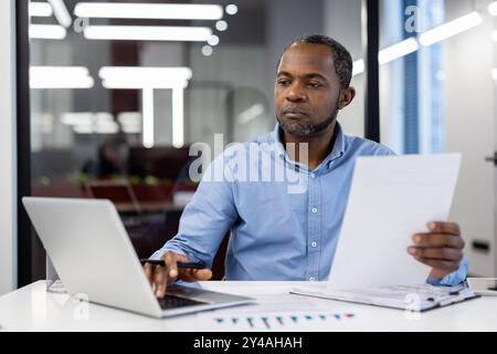 afroamerikaner reifer Geschäftsmann, der sich auf die Arbeit am Schreibtisch konzentriert. Papiere halten, Laptop im modernen Büro benutzen. Konzentration, Engagement und Professionalität im Geschäftsumfeld demonstrieren. Stockfoto