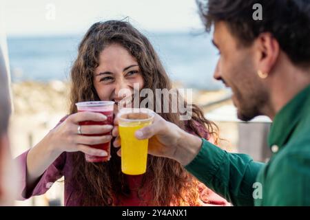 Nahaufnahme eines glücklichen jungen Paares, das einen fröhlichen Moment teilnimmt und mit lebhaften Getränken in einem Café am Strand antost. Freimütiger Schuss, der echtes Lachen einfängt Stockfoto