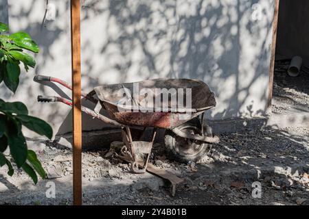 Eine verwitterte rote Schubkarre sitzt auf einem Kiesboden, teilweise von Bäumen beschattet. Die Schubkarre ist mit Schmutz und Werkzeugen gefüllt, mit einer Wand im bac Stockfoto