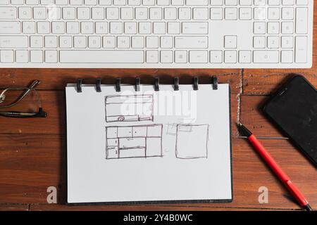Ein Skizzenbuch mit handgezeichneten Motiven, einschließlich eines Layouts mit Fernseher und Möbeln, auf einem Holztisch neben einem roten Stift und einer Computertastatur. Stockfoto