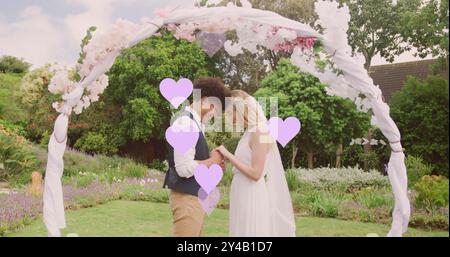 Bild von lilafarbenen Herzen und glücklichen, vielfältigen Paaren, die Hände unter dem Hochzeitsbogen im Garten halten Stockfoto