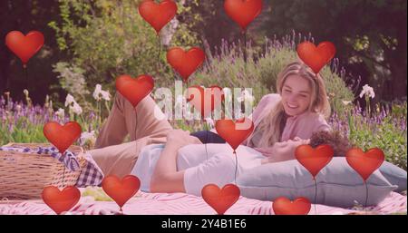Bild von roten Herzballons über glücklichen, vielfältigen Paaren, die sich beim Picknick im sonnigen Garten entspannen Stockfoto