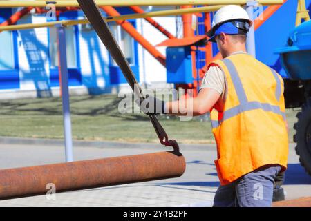 Bauarbeiter in Sicherheitsausrüstung, die ein langes Metallrohr mit einem Metallhaken auf einer Baustelle im Freien anheben und dabei die sorgfältige Handhabung und Sicherung von Baumaterialien demonstrieren. Stockfoto