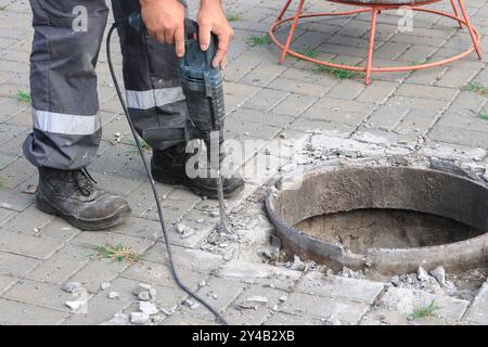 Arbeiter in Schutzausrüstung brechen mit einer Bohrmaschine Beton um eine kreisförmige Öffnung im Gehweg, um Sicherheit und Präzision auf der Baustelle zu gewährleisten. Stockfoto