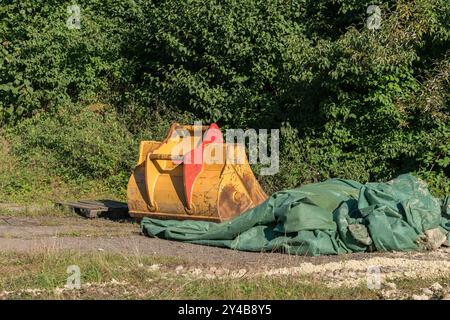 Ein gelber Baubaggerlöffel befindet sich neben einer großen grünen Plane auf einem Kiesboden, umgeben von dichtem Grün unter hellem Tageslicht Stockfoto