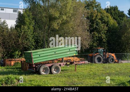 Ein grüner Anhänger sitzt im Leerlauf auf einem pulsierenden Feld neben einem Traktor, umgeben von Bäumen unter einem klaren blauen Himmel, was eine friedliche ländliche Umgebung veranschaulicht Stockfoto