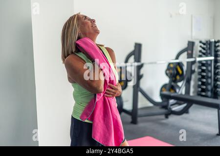 Copyspace reife blonde Frau in Sportkleidung wischt den Schweiß mit einem rosa Handtuch ab, nachdem sie eine Fitnessroutine im Fitnessstudio absolviert hat. Sie wirkt erfrischt und Stockfoto