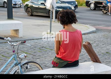 Rückansicht eines jungen afroamerikanischen Mannes mit lockigen Haaren, der neben ihrem Fahrrad auf einer Kopfsteinpflasterstraße sitzt. Städtischer Lebensstil und nachhaltiger Verkehr Stockfoto