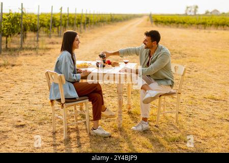 Ein paar entspannen sich an einem rustikalen Tisch in einem sonnigen Weinberg, genießen Wein und Gourmet-Snacks. Sie teilen Lachen und Freude, umgeben von üppigen Weinstöcken und dem Stockfoto