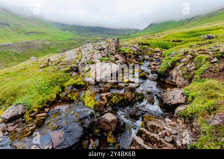 Von den schneebedeckten Hängen der Berge rund um Borgarfiordur Eystri im Osten Islands fließt ein Bach zwischen den vulkanischen Felsen Stockfoto