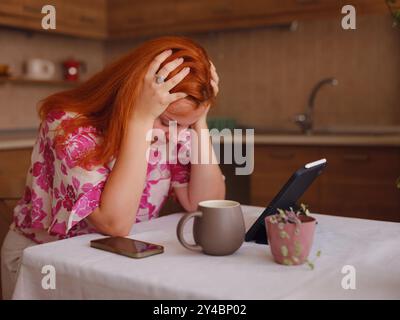 Junge Frau, die in der Küche zu Hause an einem Tablet arbeitet. Mit Kopfschmerzen am Tisch sitzen. Stress haben Probleme am Arbeitsplatz mit Blick auf Laptop-Computer i Stockfoto
