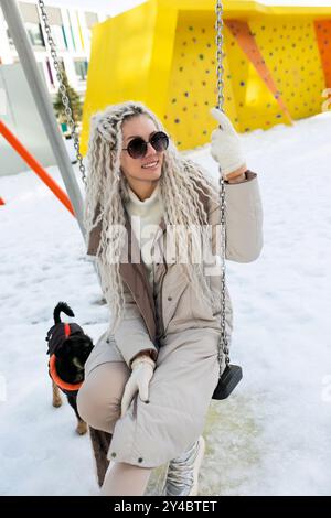 Eine Frau sitzt auf einer schneebedeckten Schaukel, gebündelt in warmer Kleidung. Der schneebedeckte Boden steht im Kontrast zu ihrem dunklen Outfit, während sie besinnlich aussieht Stockfoto