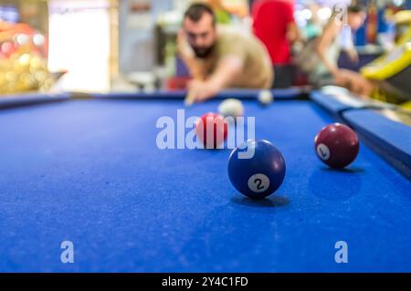 Ein fokussierter Mann, der Billard auf einem blauen Tisch spielt, umgeben von bunten Bällen in einem lebhaften und lebhaften Spielzimmer. Stockfoto