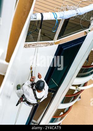 Ein Arbeiter in Bergsteigerausrüstung lackiert den Rumpf eines weißen Schiffes mit einer Malwalze in einer Höhe, ein Arbeiter in Industriebergsteiger in Uniform Stockfoto