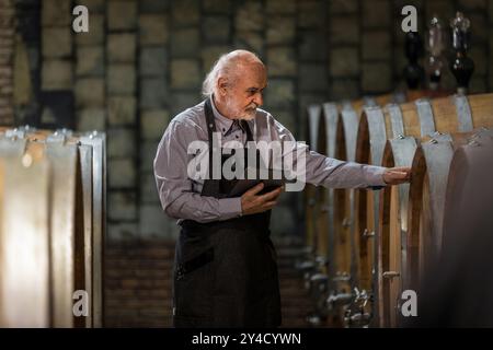 Ein im Kaukasus erfahrener Winzer überprüft eine Weinalterung in Fässern mit einem Touchpad-Tablet. Traditionelles und modernes Technologiekonzept. Stockfoto