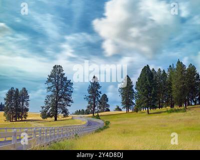 An einem hellen, teilweise bewölkten Tag im Osten Washingtons schlängelt sich eine umzäunte Straße durch das Land. Stockfoto