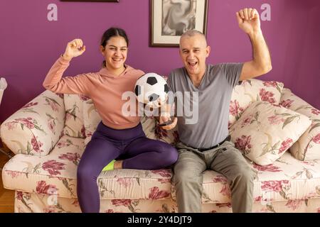 Vater und seine Tochter schauen zu Hause Fußball im Fernsehen. Emotionaler Mann und Teenager-Mädchen jubeln ihr Lieblingsteam an, Familienbegeisterung Stockfoto