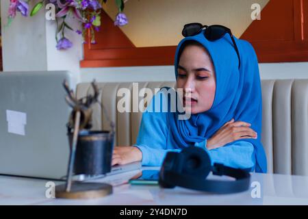 Junge asiatische Studentin, die Hijab trägt, mit Schmerzen mit Schulterschmerzen aufgrund von Müdigkeit, die Aufgaben erledigt und auf dem Sofa im Indoor Coffee Shop sitzt. Für Adver Stockfoto