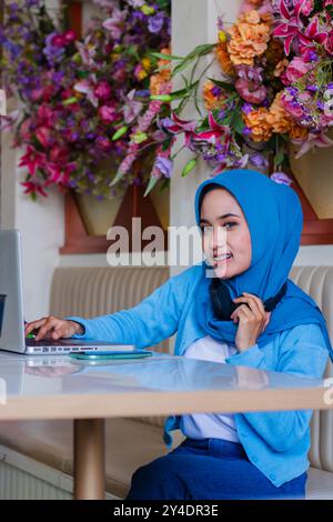 Porträt eines schönen jungen asiatischen Mädchens, das Schleier trägt, lächelt in die Kamera, indem er Daten in den Laptop eingibt, während der Kopfhörer um den Hals in einem Café im Innenbereich liegt. Für einen Stockfoto