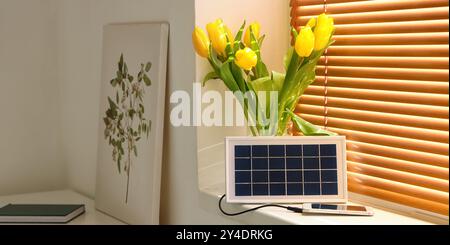 Tragbares Solarmodul-Ladegerät mit Mobiltelefon und Vase mit Tulpen auf der Fensterbank im Zimmer Stockfoto