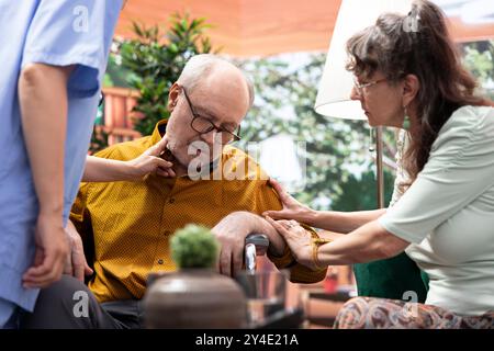 Schwester springt, um älteren Mann im Rollstuhl zu helfen, das Gewissen beim Hausbesuch zu verlieren, alte Person hyperventiliert und seine Frau erschreckt. Arzthelferin in Panikprüfung des Pulses. Stockfoto